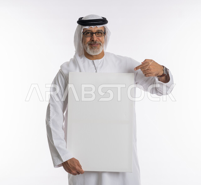لوح إعلاني فارغ، إيماءات الإعجاب والسرور والنظر إلى الكاميرا ،موك اب مستطيل أبيض ، بورتريه لرجل مسن عربي اماراتي خليجي يرتدي الكندورة و الغترة يشير باصبع السبابة الى لوحه بيضاء مستطيلة، الوقوف باستقامه وثقه، خلفية بيضاء