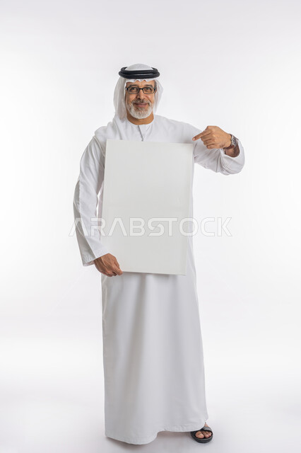 Gestures of admiration, pleasure, and looking at the camera, a white rectangular mock-up, a portrait of an elderly Arab, Emirati, Gulf man wearing a kandura and a ghutra, pointing with his index finger at a white rectangular board, an empty advertising board, standing upright and confident, full-length body portrait, white background