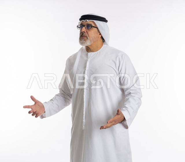 ايماءات عدم المعرفة والاستغراب، التساؤل والاستفهام والاستفسار عن شيء ما، بورتريه لرجل كبير السن عربي خليجي اماراتي يرتدي الكندورة و الغترة والنظارات الطبية يرفع يديه للاعلى بتعابير الدهشة, خلفية بيضاء