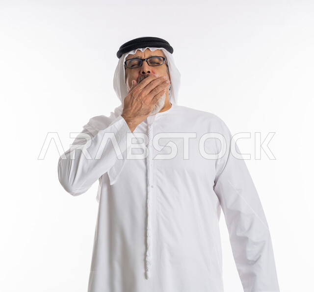الإحساس بالإرهاق و الحاجه الى النوم والراحه ,الشعور بالتعب والتثاؤب و الكسل والخمول، بورتريه مقرب لرجل كبير السن عربي خليجي اماراتي يرتدي الكندورة والغترة والنظارات الطبية يضع يده على فمه بإيماءات تدل على النعاس, خلفية بيضاء