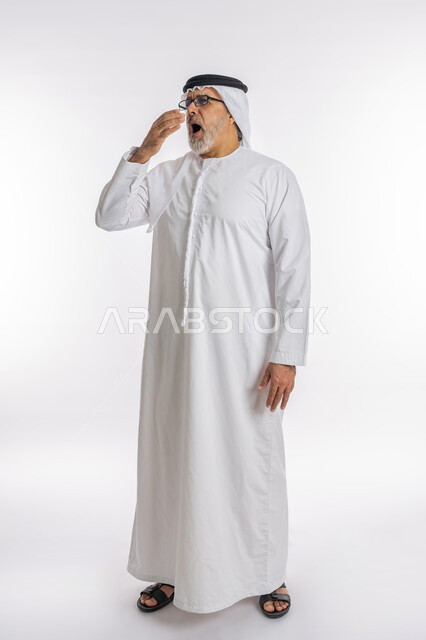 التثاؤب والشعور بالتعب و الكسل والخمول، الإحساس بالإرهاق و الحاجه الى النوم والراحه ,بورتريه لرجل كبير السن عربي خليجي اماراتي يرتدي الكندورة والغترة والنظارات الطبية يضع يده على فمه بإيماءات تدل على النعاس, الطول الكامل للجسم, خلفية بيضاء