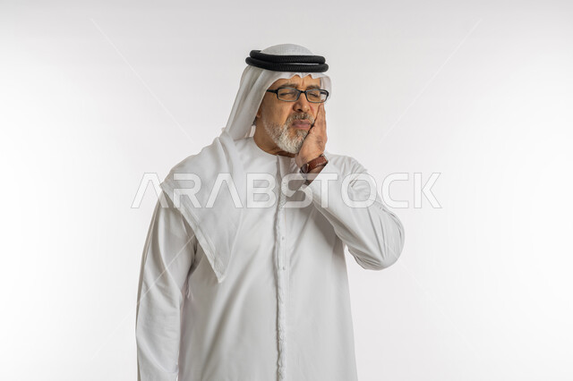 الحاجه لزيارة الطبيب، طلب العلاج والمساعده، تسوس الأضراس وإلتهاب اللثه، بورتريه مقرب لرجل كبير السن عربي خليجي اماراتي يرتدي الكندورة و الغترة و النظارات الطبيه يضع يده على خده بإيماءات تدل على الشعور بألم الأسنان، خلفية بيضاء