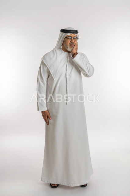 طلب العلاج والمساعده، تسوس الأضراس وإلتهاب اللثه، بورتريه لرجل كبير السن عربي خليجي اماراتي يرتدي الكندورة و الغترة و النظارات الطبيه يضع يده على خده بإيماءات تدل على الشعور بألم الأسنان، الحاجه لزيارة الطبيب، الجسم بالطول الكامل, خلفية بيضاء