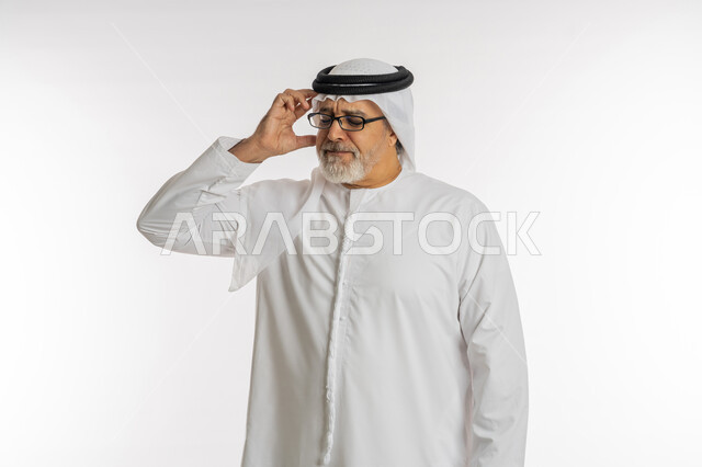 آلام الشقيقة والصداع النصفي ,الإحساس بوجع الرأس و الدوخة ,الحاجة للعلاج والراحة ,إيماءات الألم والتعب والمرض, بورتريه مقرب لرجل كبير السن عربي خليجي اماراتي يرتدي الغترة والكندورة و النظارات الطبية يضع يديه على رأسه يشعر بالدوار, خلفية بيضاء