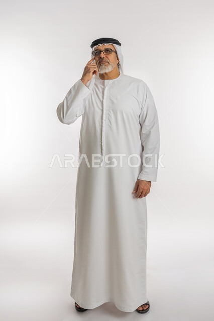 Absent-mindedness gestures, making decisions with concentration, trying to find a solution to problems, looking upward carefully and planning for the future, portrait of an old Arab Gulf Emirati man wearing a ghutra, kandura and medical glasses, placing his hand on his face with a thinking expression, full-length body, white background