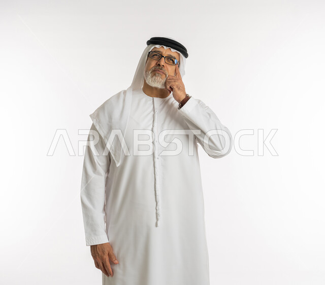 Looking up carefully and planning for the future, absent-mindedness and focused decision-making, trying to find a solution to problems, portrait of an elderly Arab, Gulf, Emirati man wearing a ghutra, kandura, and medical glasses, placing his hand on his face with a thinking expression, white background.