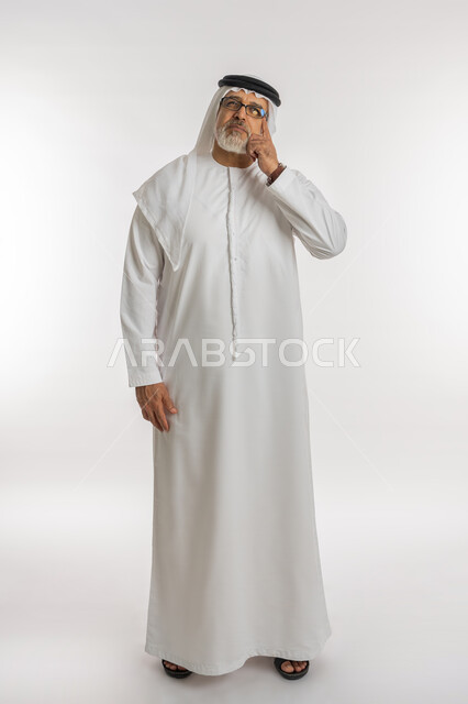 Absent-mindedness and focused decision-making, trying to find a solution to problems, looking carefully and planning for the future, portrait of an elderly Arab, Gulf, Emirati man wearing a ghutra, kandora, and medical glasses, placing his hand on his face with a thinking expression, full-length body, white background