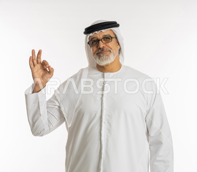 إيماءات علامة الجودة و السرور, النظر إلى الكاميرا بإشارات الموافقه, الشعور بالرضا و السعادة, بورتريه مقرب لرجل كبير السن عربي خليجي اماراتي مبتسم يرتدي الكندورة و الغترة يرفع يده للأعلى بحركات الاعجاب والقبول, خلفية بيضاء