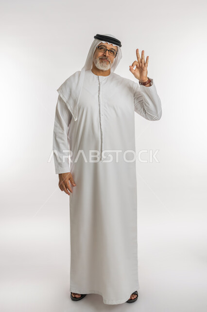 حركات الاعجاب والقبول, الشعور بالرضا و السعادة, النظر إلى الكاميرا بإشارات الموافقه، بورتريه لرجل كبير السن عربي خليجي اماراتي مبتسم يرتدي الكندورة و الغترة يرفع يده للأعلى بإيماءات علامة الجودة و السرور, صورة الجسم بالطول الكامل، خلفية بيضاء