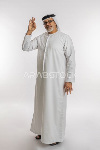 الشعور بالرضا و السعادة, النظر إلى الكاميرا بإشارات الموافقه، حركات الاعجاب والقبول, بورتريه لرجل كبير السن عربي خليجي اماراتي مبتسم يرتدي الكندورة و الغترة يرفع يده للأعلى بإيماءات علامة الجودة و السرور, صورة الجسم بالطول الكامل، خلفية بيضاء