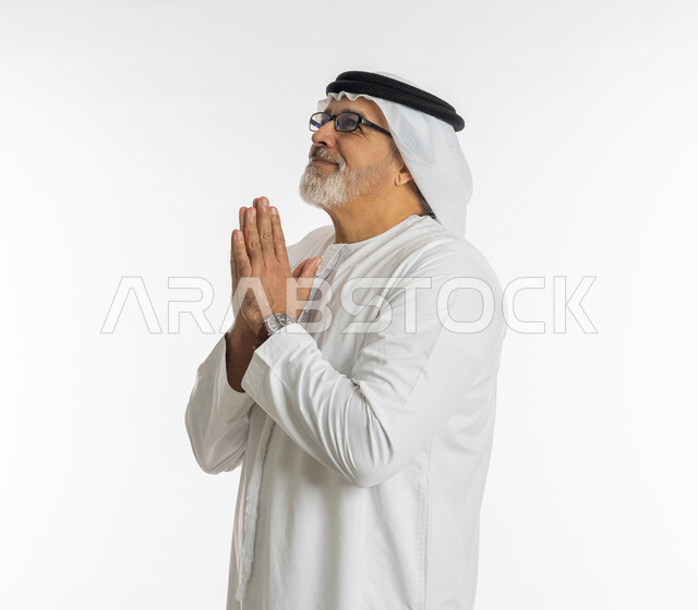مفهوم طلب المغفرة و العون، الرغبة في تحقيق الأمنيات, الحمد والشكر لله، بورتريه مقرب لرجل كبير السن عربي اماراتي خليجي يرتدي الغترة والكندورة و النظارات الطبية يرفع يديه بايماءات الدعاء والتضرع، خلفية بيضاء