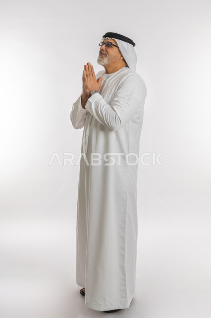 مفهوم طلب العون و المغفرة، الحمد والشكر لله، بورتريه لرجل كبير السن عربي اماراتي خليجي يرتدي الغترة والكندورة و النظارات الطبية يرفع يديه بايماءات الدعاء والتضرع، الرغبة في تحقيق الأمنيات، الجسم بالطول الكامل, خلفية بيضاء