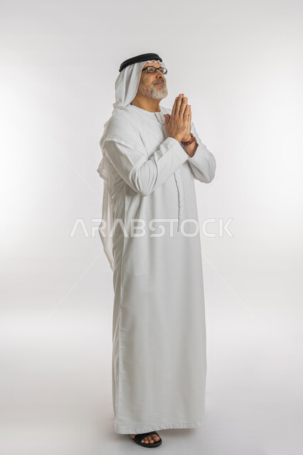 الحمد والشكر لله، بورتريه لرجل كبير السن عربي اماراتي خليجي يرتدي الغترة والكندورة و النظارات الطبية يرفع يديه بايماءات الدعاء والتضرع، مفهوم طلب العون و المغفرة، الرغبة في تحقيق الأمنيات، الجسم بالطول الكامل, خلفية بيضاء