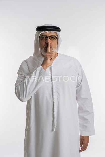 الأمر بالسكوت والصمت، التوقف عن الكلام وخفض الصوت، بورتريه من الجانب لرجل كبير السن عربي خليجي اماراتي يرتدي الكندورة و الغترة و النظارات الطبية يقف باستقامة و يضع أصبع السبابة على فمه بإيماءات طلب عدم الإزعاج و السكون والهدوء ,خلفية بيضاء