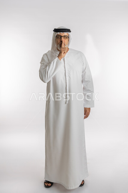 الأمر بالصمت و السكوت، التوقف عن الكلام وخفض الصوت، بورتريه من الجانب لرجل كبير السن عربي خليجي اماراتي يرتدي الكندورة و الغترة و النظارات الطبية يقف باستقامة و يضع أصبع السبابة على فمه بإيماءات طلب عدم الإزعاج و السكون والهدوء, الجسم بالطول الكامل, خلفية بيضاء