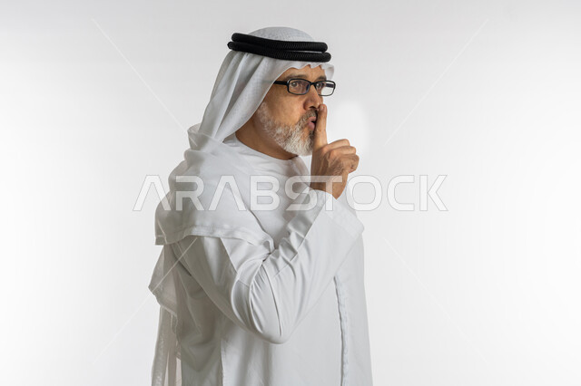 عدم الإزعاج و السكون والهدوء، التوقف عن الكلام وخفض الصوت، بورتريه مقرب من الجانب لرجل كبير السن عربي خليجي اماراتي يرتدي الكندورة و الغترة و النظارات الطبية يضع أصبع السبابة على فمه بإيماءات طلب السكوت والصمت, خلفية بيضاء