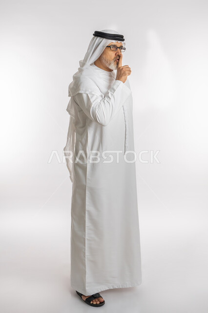 السكون والهدوء و عدم الإزعاج، التوقف عن الكلام وخفض الصوت، بورتريه من الجانب لرجل كبير السن عربي خليجي اماراتي يرتدي الكندورة و الغترة و النظارات الطبية يضع أصبع السبابة على فمه بإيماءات طلب السكوت والصمت، صورة الجسم بالطول الكامل, خلفية بيضاء