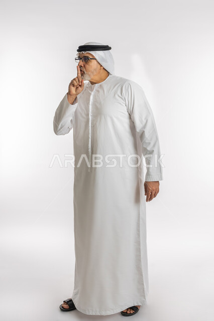 الهدوء والسكون وعدم الإزعاج، التوقف عن الكلام وخفض الصوت، بورتريه لرجل كبير السن عربي خليجي اماراتي يرتدي الكندورة و الغترة و النظارات الطبية يضع أصبع السبابة على فمه بإيماءات طلب السكوت والصمت، صورة الجسم بالطول الكامل, خلفية بيضاء