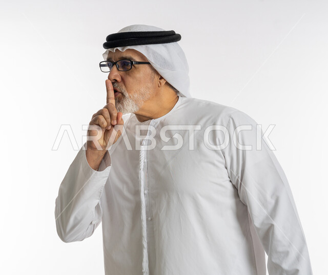 التوقف عن الكلام وخفض الصوت، بورتريه مقرب لرجل كبير السن عربي خليجي اماراتي يرتدي الكندورة و الغترة و النظارات الطبية يضع أصبع السبابة على فمه بإيماءات طلب السكوت والصمت، الهدوء والسكون وعدم الإزعاج، خلفية بيضاء