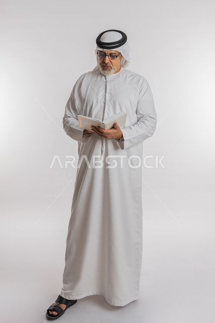 حب الاستكشاف و المطالعة ,مفهوم محو الامية، تعابير التركيز أثناء القراءة، قضاء وقت الفراغ بالتعلم والثقافة، بورتريه لرجل كبير بالسن عربي اماراتي خليجي يرتدي الكندورة و الغترة و النظارات الطبية يحمل بيده كتاب علمي تثقيفي، الجسم بالطول الكامل, خلفية بيضاء