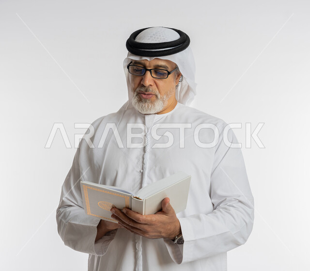 مفهوم محو الامية، تعابير التركيز أثناء القراءة، هواية حب المطالعة و الاستكشاف, قضاء وقت الفراغ بالتعلم والثقافة، بورتريه مقرب لرجل كبير بالسن عربي اماراتي خليجي يرتدي الكندورة و الغترة و النظارات الطبية يحمل بيده كتاب علمي تثقيفي، خلفية بيضاء