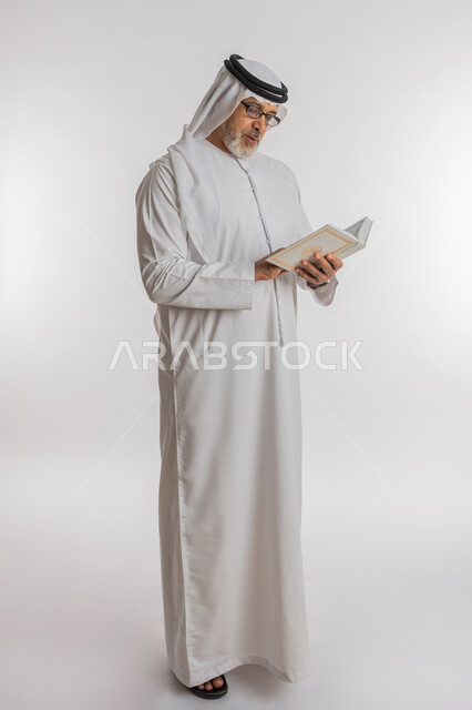 تعابير الاندماج و التركيز اثناء القراءة، حب المطالعة و الاستكشاف, قضاء وقت الفراغ بالتعلم ، مفهوم محو الامية، بورتريه لرجل كبير بالسن عربي اماراتي خليجي يرتدي الكندورة و الغترة و النظارات الطبية يحمل بيده كتاب علمي تثقيفي، الطول الكامل للجسم, خلفية بيضاء