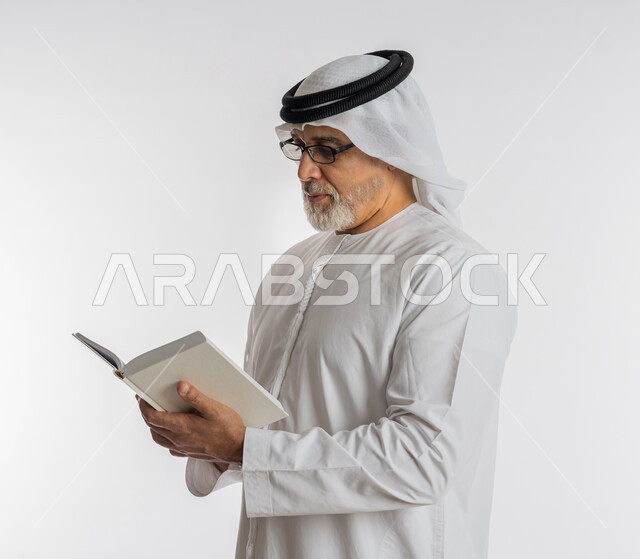 القراءة بتركيز وتمعن، هواية حب المطالعة و الاستكشاف, قضاء وقت الفراغ بالتعلم والثقافة، مفهوم محو الامية، بورتريه مقرب لرجل كبير بالسن عربي اماراتي خليجي يرتدي الكندورة و الغترة و النظارات الطبية يحمل بيده كتاب علمي تثقيفي، خلفية بيضاء