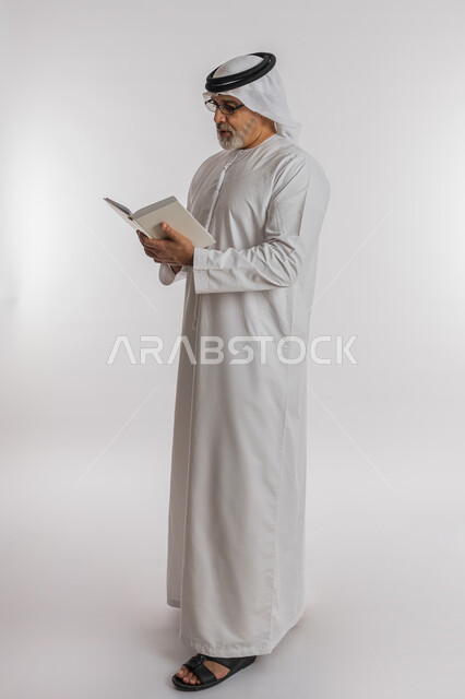 قضاء وقت الفراغ بالتعلم والثقافة، حب المطالعة و الاستكشاف ,القراءة بتركيز وتمعن، مفهوم محو الامية، بورتريه لرجل كبير بالسن عربي اماراتي خليجي مبتسم يرتدي الكندورة و الغترة و النظارات الطبية يحمل بيده كتاب علمي تثقيفي، صورة الجسم بالطول الكامل, خلفية بيضاء