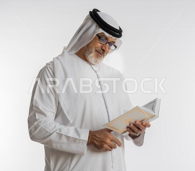 حب المطالعة و الاستكشاف ,القراءة بتركيز وتمعن، قضاء وقت الفراغ بالتعلم والثقافة، مفهوم محو الامية، بورتريه مقرب لرجل كبير بالسن عربي اماراتي خليجي مبتسم يرتدي الكندورة و الغترة و النظارات الطبية يحمل بيده كتاب علمي تثقيفي، خلفية بيضاء