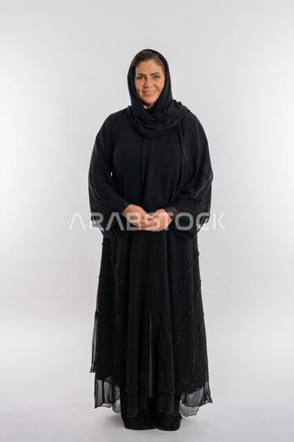 The concept of elegance and attention to external appearance and femininity, a portrait of a veiled Saudi Arab woman wearing a black abaya looking at the camera with an expression of pleasure, a smiling Emirati Gulf woman standing upright with gestures of self-confidence, full-length body portrait, white background