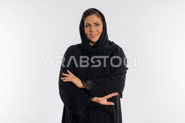 The concept of elegance and femininity, looking at the camera with expressions of self-confidence, a close-up portrait of a veiled Saudi Arab woman wearing a black abaya, standing with shoulder-to-shoulder hand movements, a smiling Gulf Emirati woman placing her palms on her arms with gestures of pleasure, white background