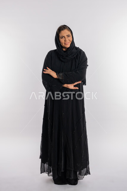 مفهوم الاهتمام بالمظهر الخارجي ,النظر الى الكاميرا بتعابير الثقة بالنفس, بورتريه لسيدة عربيه سعودية محجبة ترتدي العباءه السوداء تقف بحركات أيدي متكتفة, امرأة خليجية اماراتية مبتسمة تضع كفيها على ذراعيها بايماءات السرور, الجسم بالطول الكامل, خلفية بيضاء