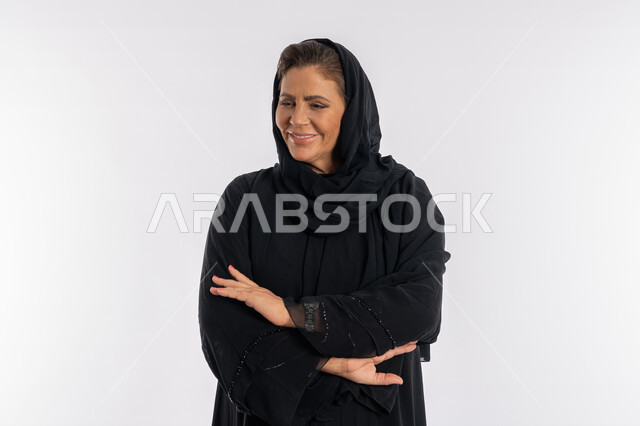 Modesty, softness and elegance, looking at the camera with an expression of shyness, a close-up portrait of a veiled Saudi Arab woman wearing a black abaya, standing with folded hand movements, attention to external appearance, a smiling Emirati Gulf woman placing her palms on her arms with gestures of pleasure, white background