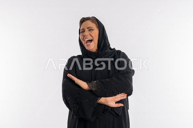 الوقوف بحركات أيدي متكتفة, بورتريه مقرب لسيدة عربيه سعودية محجبة ترتدي العباءه السوداء تضحك بطريقة هستيرية, الاهتمام بالمظهر الخارجي ,امرأة خليجية اماراتية مسرورة تضع كفيها على ذراعيها بايماءات الفرح, خلفية بيضاء