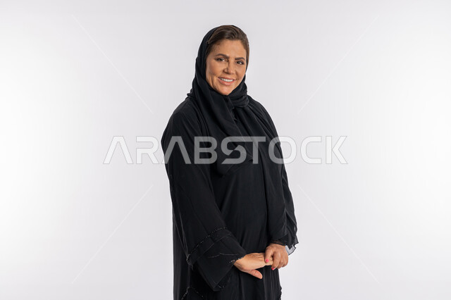 مفهوم الخجل و النعومة و الأناقة ,الاهتمام بالمظهر الخارجي، بورتريه لامرأة عربية إماراتية محجبة ترتدي العباءة السوداء وتنظر الى الكاميرا بإيماءات الحياء، سيدة خليجية سعودية مبتسمة تقف بتعابير أيدي متماسكة، خلفية بيضاء