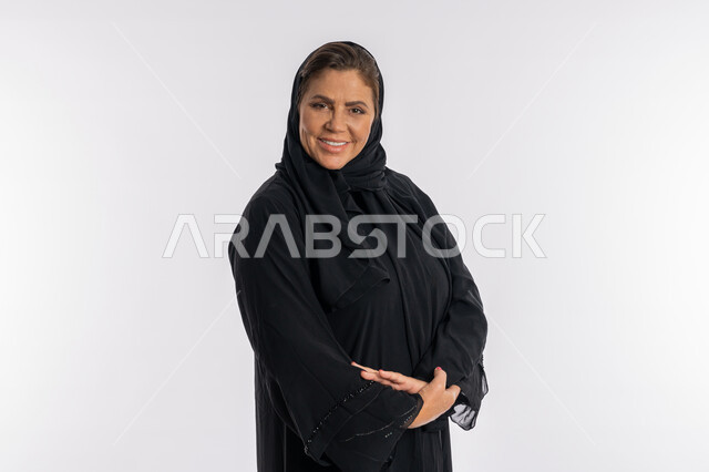 الوقوف بحركات أيدي متكتفة, بورتريه مقرب لسيدة عربيه سعودية محجبة ترتدي العباءه السوداء تنظر الى الكاميرا بتعابير الفرح, الاهتمام بالمظهر الخارجي ,امرأة خليجية اماراتية مبتسمة تضع كفيها على ذراعيها بايماءات السرور, خلفية بيضاء