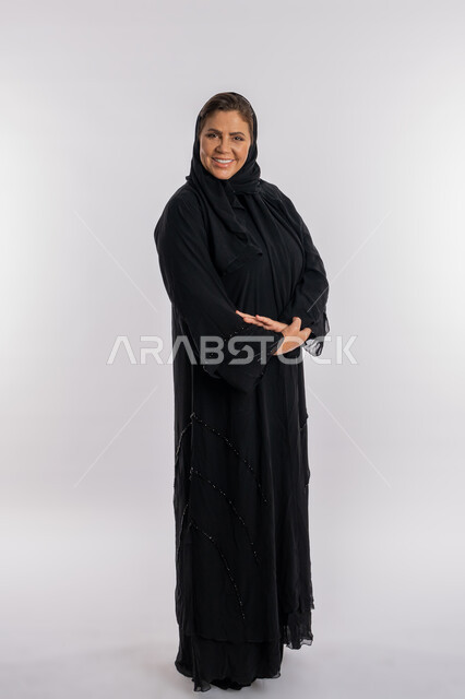 الاهتمام بالمظهر الخارجي ,الوقوف بحركات أيدي متكتفة, بورتريه لسيدة عربيه سعودية محجبة ترتدي العباءه السوداء تنظر الى الكاميرا بتعابير الفرح, امرأة خليجية اماراتية مبتسمة تضع كفيها على ذراعيها بايماءات السرور, صورة الجسم بالطول الكامل, خلفية بيضاء
