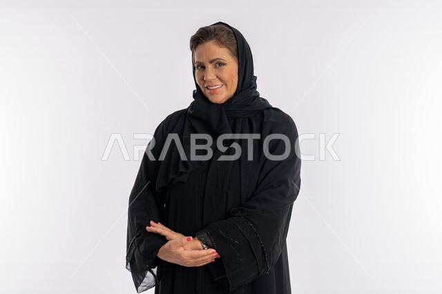 النظر الى الكاميرا بتعابير الفرح, بورتريه مقرب لسيدة عربيه سعودية محجبة ترتدي العباءه السوداء تقف بحركات أيدي متكتفة, الاهتمام بالمظهر الخارجي ,امرأة خليجية اماراتية مبتسمة تضع كفيها على ذراعيها بايماءات السرور, خلفية بيضاء