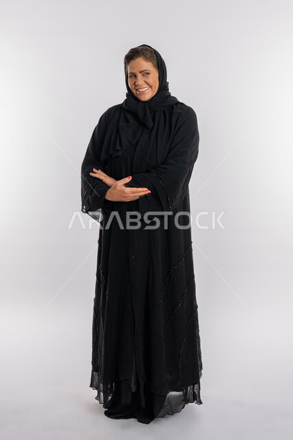 الوقوف بحركات أيدي متكتفة, بورتريه لسيدة عربيه سعودية محجبة ترتدي العباءه السوداء تنظر الى الكاميرا بتعابير الفرح, امرأة خليجية اماراتية مبتسمة تضع كفيها على ذراعيها بايماءات السرور, صورة الجسم بالطول الكامل, خلفية بيضاء