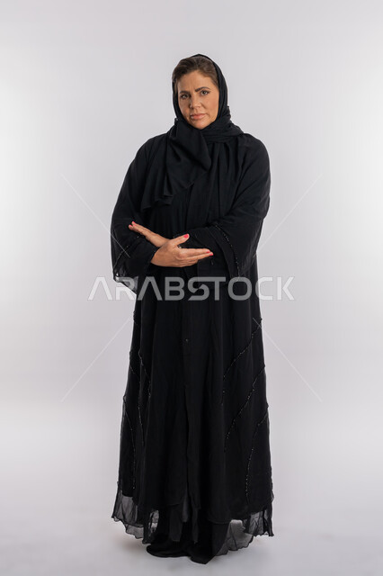النظر الى الكاميرا بحركات الجدية, بورتريه لسيدة عربيه سعودية محجبة ترتدي العباءه السوداء تقف بتعابير أيدي متكتفة, امرأة خليجية اماراتية تضع كفيها على ذراعيها بايماءات الثقة بالنفس, صورة الجسم بالطول الكامل, خلفية بيضاء