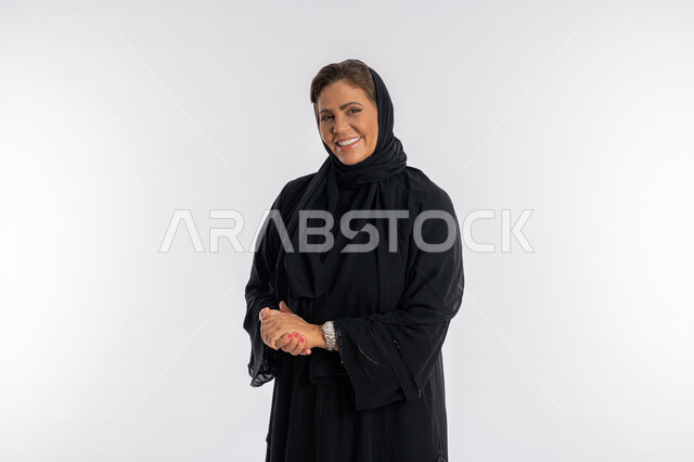 الاهتمام بالمظهر الخارجي ,مفهوم الاناقة و الأنوثة, بورتريه مقرب لامرأة عربيه سعودية محجبة ترتدي العباءه السوداء تنظر الى الكاميرا بتعابير السرور, سيدة خليجية اماراتية مبتسمة تقف باستقامة بايماءات أيدي متماسكة, خلفية بيضاء