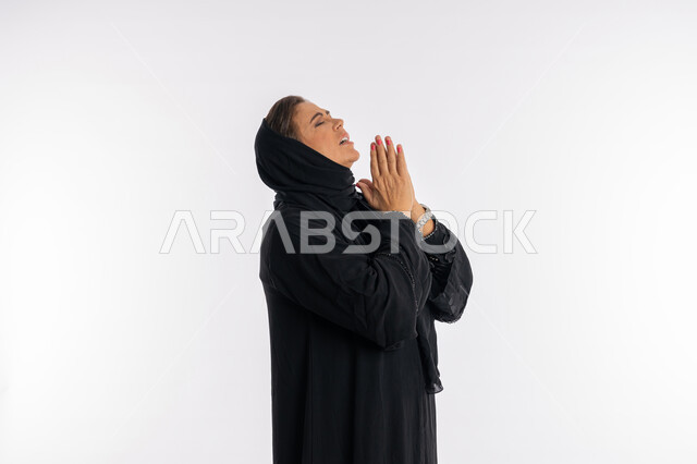 مفهوم طلب العون و المغفرة، الحمد والشكر لله، بورتريه مقرب من الجانب لامرأة عربية سعودية محجبة ترتدي العباءة السوداء ترفع يديها بايماءات الدعاء والتضرع، سيدة اماراتية خليجية ترغب في تحقيق الأمنيات، خلفية بيضاء