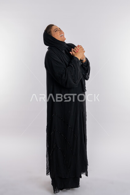 الوقوف بحركات أيدي متماسكة بإيماءات السعادة والسرور,  بورتريه لسيدة عربية اماراتية محجبة ترتدي العباءه السوداء ترفع رأسها عاليا بتعابير الضحك الهستيري ,امراة خليجية سعودية تفرح عند سماع خبر معين, صورة الجسم بالطول الكامل, خلفية بيضاء