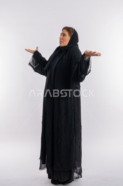 الاستغراب والاستفسار و التساؤل من أمر ما، سيدة سعودية عربية تفتح يديها بتعابير الإستفهام حول موضوع معين, الغموض وعدم الوضوح، بورتريه لامرأة خليجية اماراتية محجبة ترتدي العباءة السوداء تفتح ذراعيها بإيماءات عدم المعرفة، الجسم بالطول الكامل, خلفية بيضاء