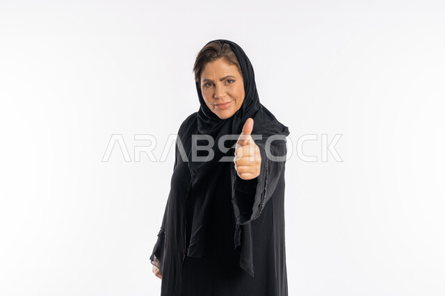 النظر الى الكاميرا بتعابير السرور، سيدة سعودية تشعر بالإعجاب، بورتريه مقرب لامراة عربية خليجية اماراتية مبتسمة محجبة ترتدي العباءة السوداء ترفع اصبع الابهام للاعلى بإيماءات علامة الجودة و الامتياز, الشعور بالرضى و القبول و الموافقة ,خلفية بيضاء