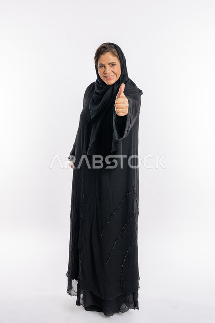 النظر الى الكاميرا بتعابير السرور, علامة الجودة و الامتياز، سيدة سعودية تشعر بالإعجاب والرضى، بورتريه لامراة عربية خليجية اماراتية مبتسمة محجبة ترتدي العباءة السوداء ترفع اصبع الابهام للاعلى بإيماءات القبول و الموافقة، الجسم بالطول الكامل, خلفية بيضاء