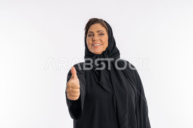 الشعور بالرضى و القبول و الموافقة ,النظر الى الكاميرا بتعابير السرور، سيدة سعودية تشعر بالإعجاب، بورتريه مقرب لامراة عربية خليجية اماراتية مبتسمة محجبة ترتدي العباءة السوداء ترفع اصبع الابهام للاعلى بإيماءات علامة الجودة و الامتياز, خلفية بيضاء