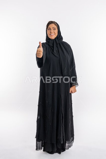 علامة الجودة و الامتياز، النظر الى الكاميرا بتعابير السرور, سيدة سعودية تشعر بالإعجاب والرضى، بورتريه لامراة عربية خليجية اماراتية مبتسمة محجبة ترتدي العباءة السوداء ترفع اصبع الابهام للاعلى بإيماءات القبول و الموافقة، الجسم بالطول الكامل, خلفية بيضاء