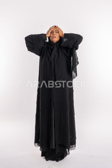 الشعور بالاحباط و الندم على شيء ما, بورتريه لإمرأة عربيه سعوديه محجبة ترتدي العباءه السوداء تضع يديها على رأسها بتعابير الدهشه و الانزعاج, الوقوع في مصيبه, سيدة اماراتية خليجية تشعر بالخسارة و الحزن, الجسم بالطول الكامل, خلفية بيضاء