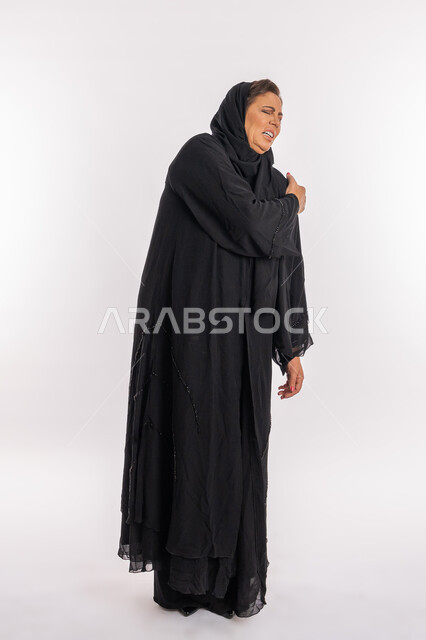 التهاب الاعصاب و العضلات والمفاصل، الحاجه للعلاج وطلب المساعده، بورتريه لامرأة خليجيه إماراتية ترتدي العباءة السوداء تشعر بالمرض و التعب، سيدة عربية سعودية تضع يدها على كتفها بإيماءات تدل على الألم الشديد، صورة الجسم بالطول الكامل, خلفية بيضاء